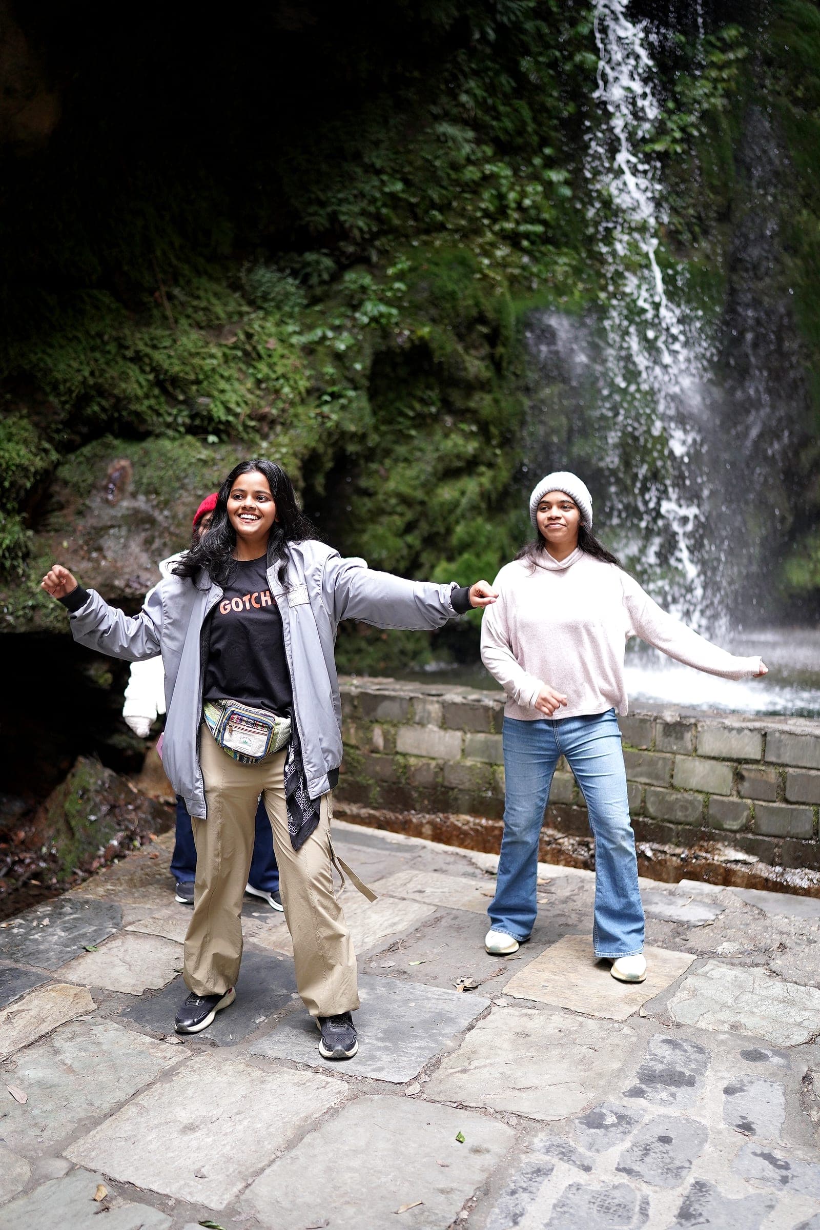 Two travellers standing arms outstretched and laughing in front of a waterfall in Himachal Pradesh